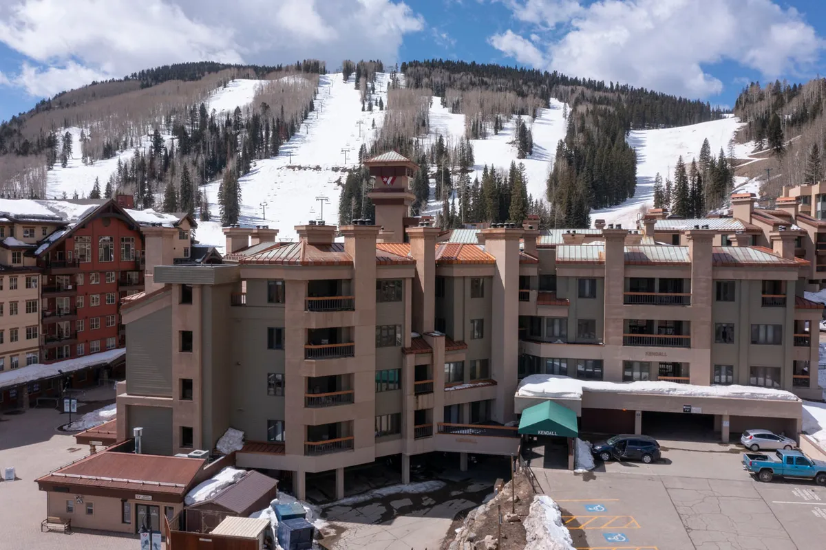 A clear exterior shot of the main resort building set against snow-covered mountain slopes. The scene reveals the lodge’s welcoming entrance, the surrounding natural landscape, and a lightly bustling parking area. The ambiance blends the thrill of alpine activity with the warmth and comfort of arriving home after a day of adventure.