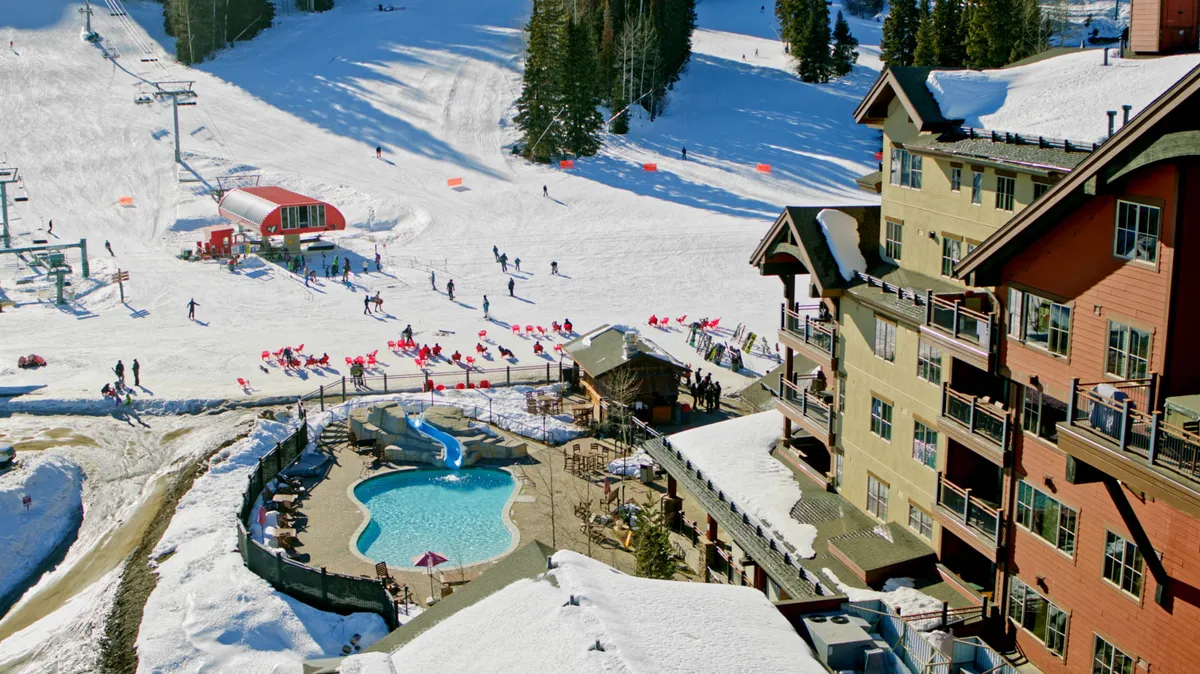 An exterior view showing the ski slopes running alongside a lively resort area, complete with a swimming pool and visible ski lifts. Resort buildings provide a charming backdrop, and people can be seen enjoying both snow activities and sunny poolside relaxation, creating a vibrant sense of community and fun.