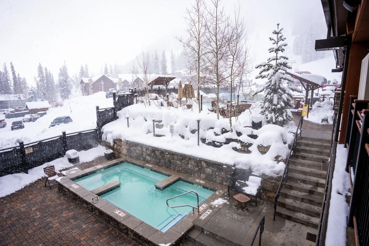 This image showcases an inviting outdoor hot tub set against a serene, snow-covered landscape, surrounded by trees and accessed by a staircase. The cozy atmosphere and glimpses of people relaxing capture the allure of soaking under the stars or watching snowflakes fall while enjoying the warmth of the hot tub.