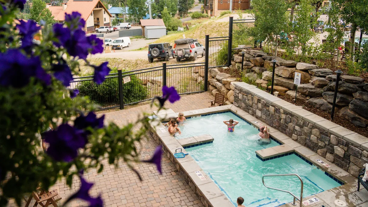 An exterior view of a heated outdoor swimming pool bordered by vibrant purple flowers, with people lounging and socializing poolside. The scene is set during sunset, offering a relaxed, welcoming mood with mountain views in the background.