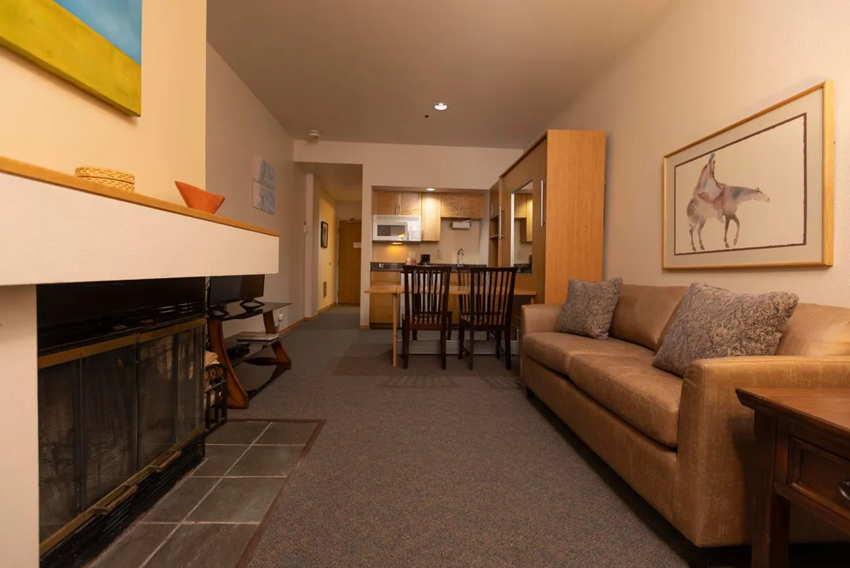 The image showcases a cozy, tastefully decorated interior with a glowing wood-burning fireplace as the centerpiece. A plush sofa is positioned for relaxing fireside evenings, while a nearby dining table invites intimate meals or celebrations. Warm lighting and inviting artwork add to the heartfelt, welcoming atmosphere.