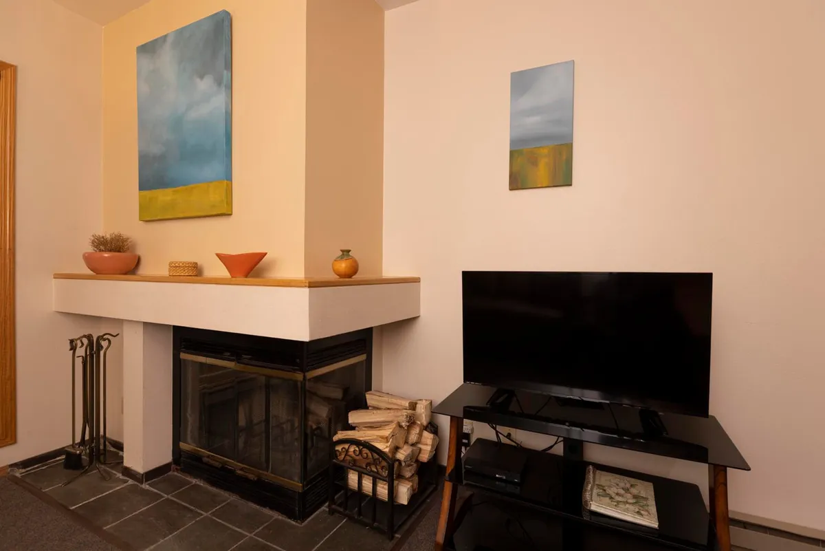 A warmly lit living area centers on a wood-burning fireplace, with wood neatly stacked nearby. Comfortable furnishings invite relaxation, and cozy décor like paintings and soft lighting create an intimate, welcoming atmosphere.