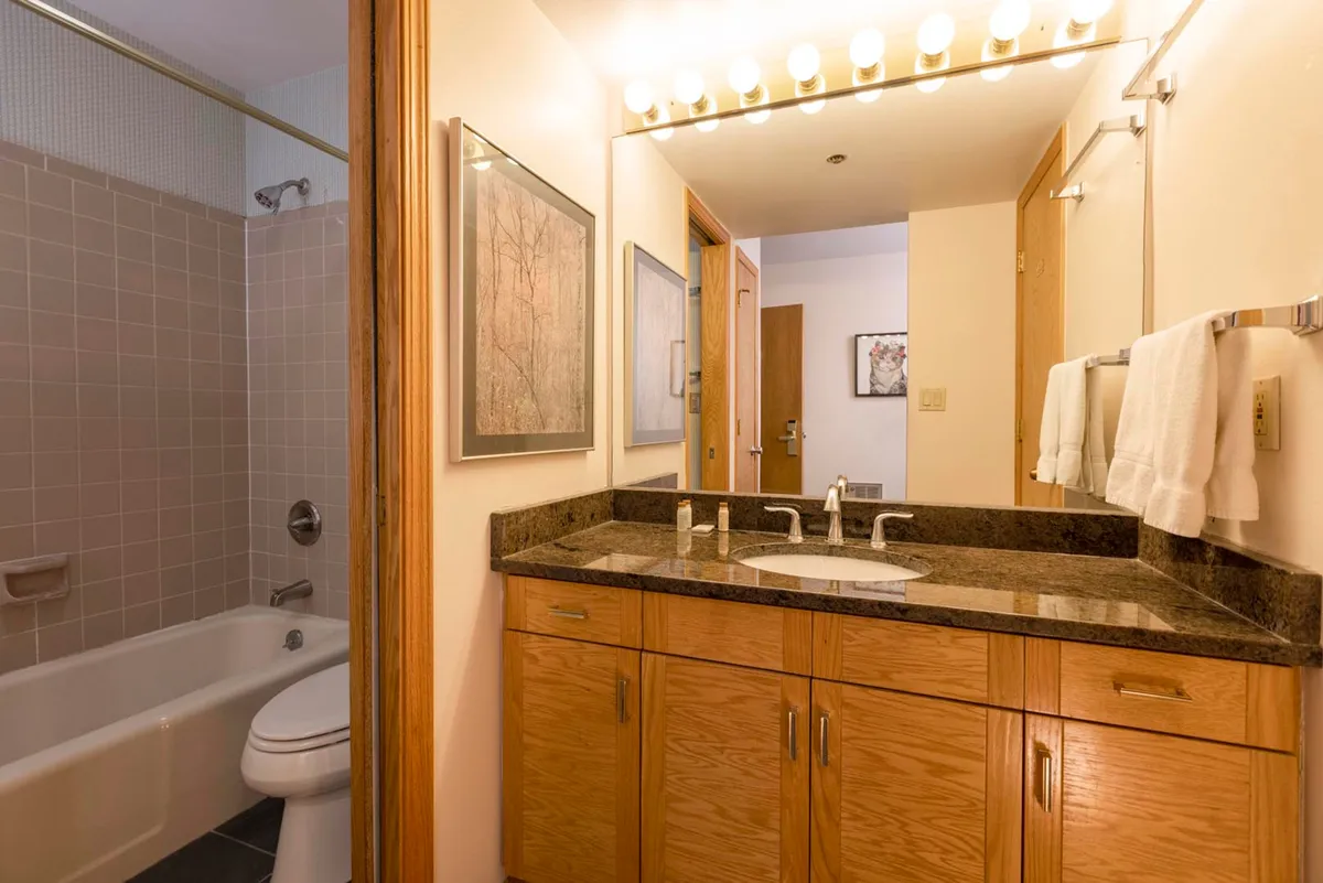 This image showcases a serene bathroom with a double vanity sink, a well-lit mirrored lighting fixture, framed artwork, and a bathtub. The space is well-appointed, inviting, and exudes a refreshing, spa-like ambiance, perfectly capturing both the practical and restorative aspects of a private ensuite bath.