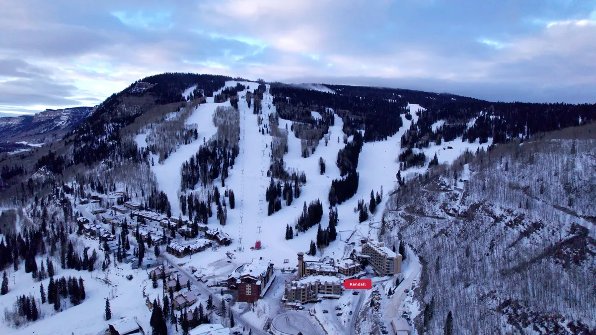 A sweeping view looking out over the ski slopes and resort buildings, with dense forest and dramatic mountain ranges in the background. The scene is alive with skiers and snowboarders, hinting at bustling activity, with the resort village nestled comfortably at the base of the mountain. The lighting suggests either a vibrant morning or the approach of dusk, lending an adventurous and inviting atmosphere.