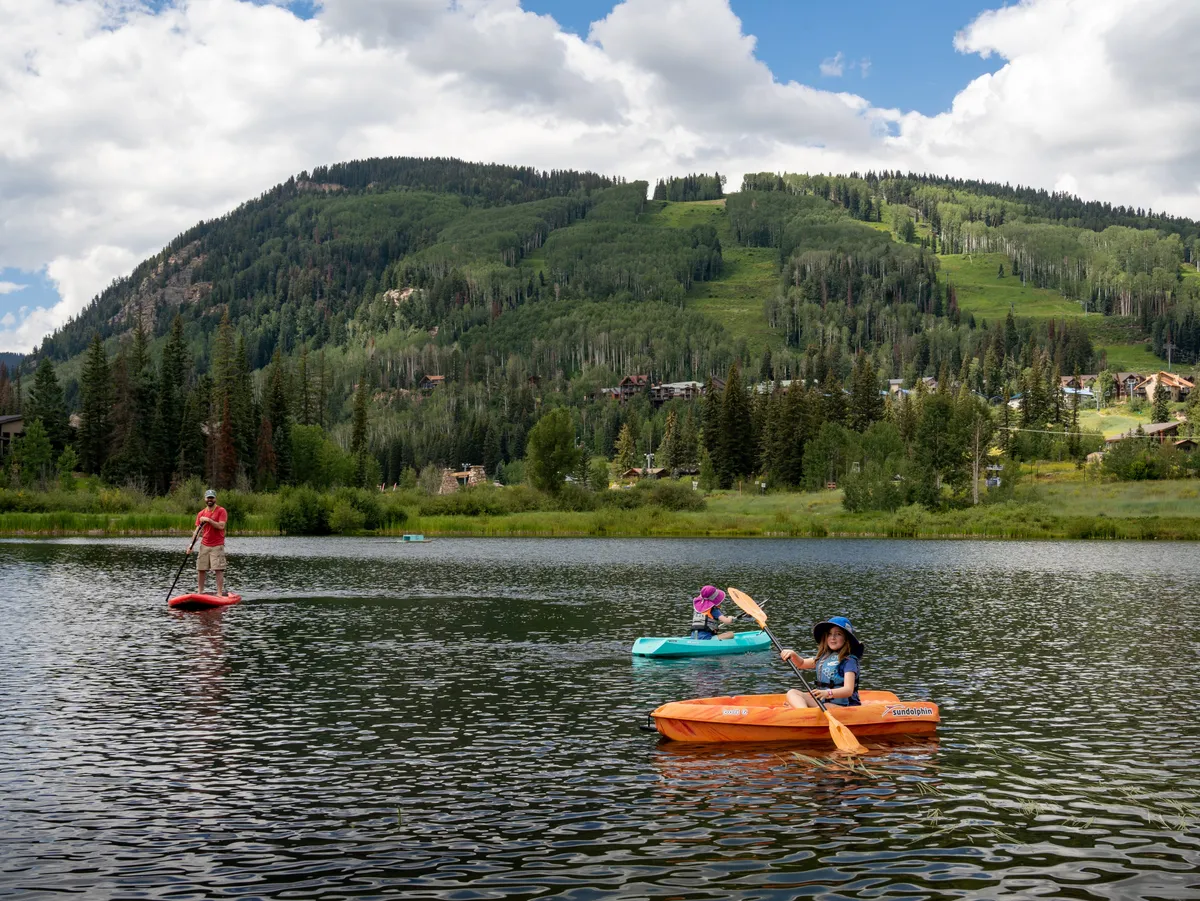 A scenic view featuring people kayaking and paddleboarding on a tranquil lake, with a dramatic mountain backdrop reflecting in the water. The natural setting is lively and inviting, hinting at the variety of leisure activities and the relaxing atmosphere that pervades the area.