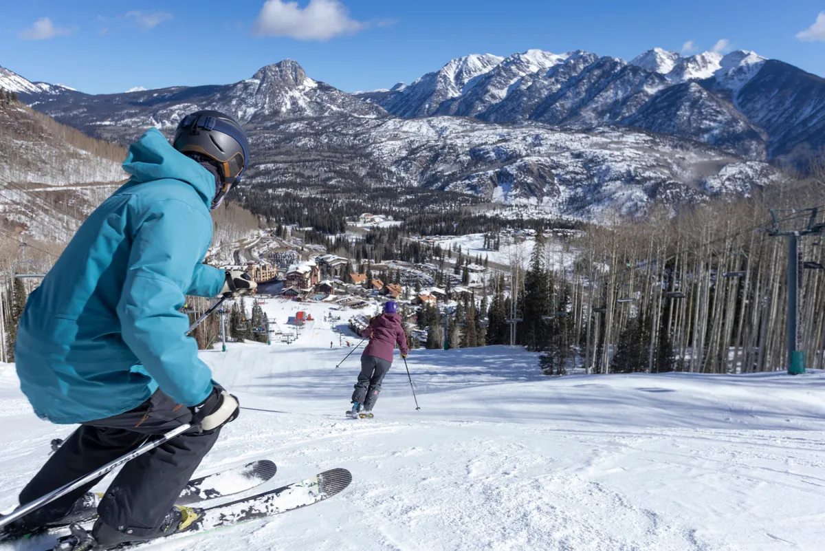 This image captures skiers descending a snowy mountain slope with the resort town visible in the valley below, set against a backdrop of majestic, snow-covered peaks. The exterior setting conveys a vibrant après-ski atmosphere and the allure of the surrounding mountain landscape.